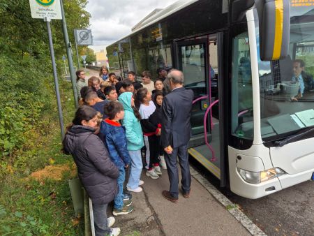 Schulkinder stehen vor einem leeren Bus und bekommen von einem Mann in einem Anzug eine Einweisung. Die Kinder hören aufmerksam zu.