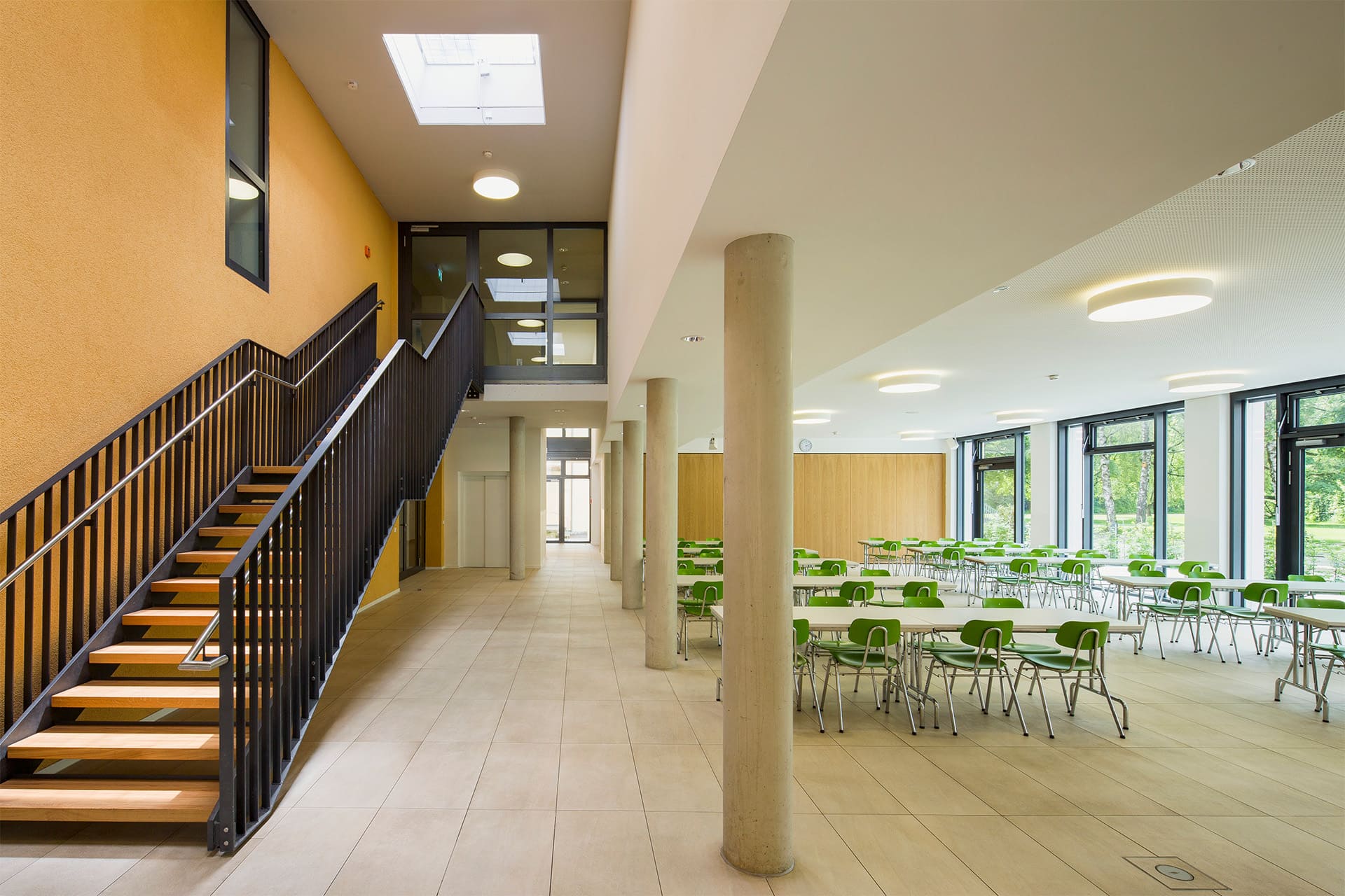 Auf der linken Seite führt eine Treppe in den 1. Stock und rechts sind die Tische und Stühle der Mensa zu sehen. Der Raum ist menschenleer.