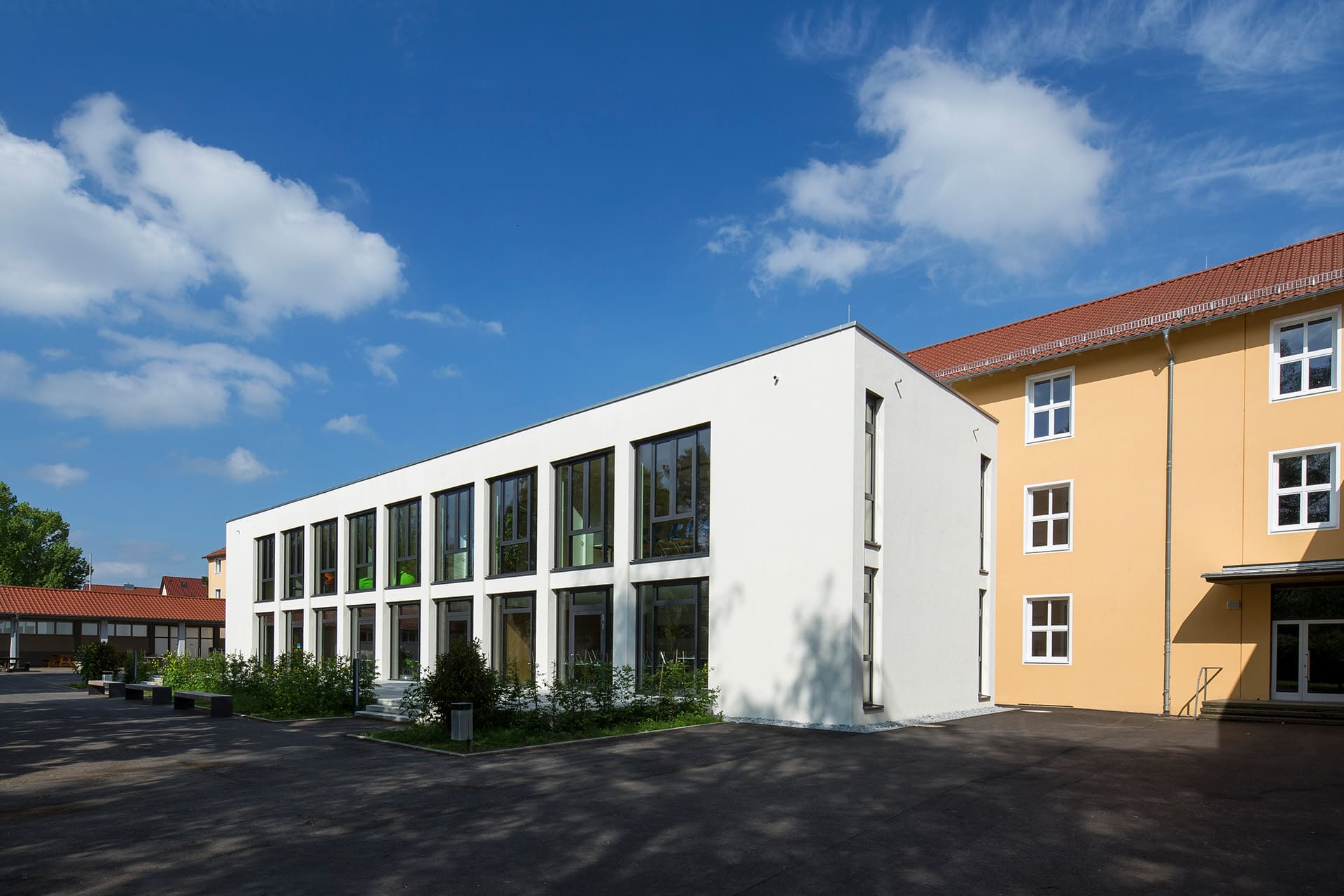 Die Mensa ist vom Schulhof zu sehen. Es ist schönes Wetter. Vor der Mensa stehen ein paar Bänke vor grünen Sträuchern.
