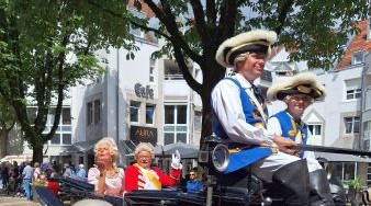 eine Frau und ein Mann altmodisch verkleidet in einem Wagen lächeln und winken