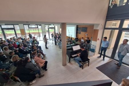 Mehrere Personen, die auf Stühlen sitzen. Vorne an einer Leinwand spielt dann ein Schulorchester.
