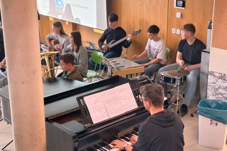 Eine Schulband, die gerade etwas vorführt. Im Vordergrund ein Mann an einem Klavier dahinter Schüler die Blas-, Seiten und Schlaginstrumente spielen