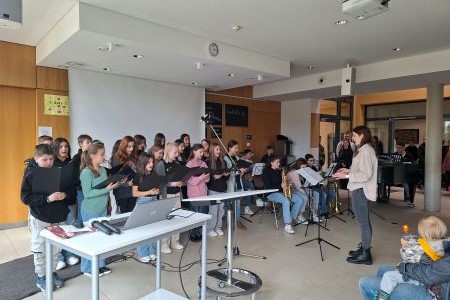 Eine Dirigentin und ein Chor, rechts daneben Personen die Instrumente spielen
