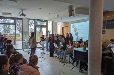 Eine Dirigentin und ein Chor, rechts daneben Personen die Instrumente spielen