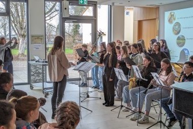 Eine Dirigentin und ein Chor, rechts daneben Personen die Instrumente spielen