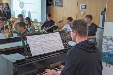 Eine Schulband, die gerade etwas vorführt. Im Vordergrund ein Mann an einem Klavier dahinter Schüler die Blas-, Seiten und Schlaginstrumente spielen